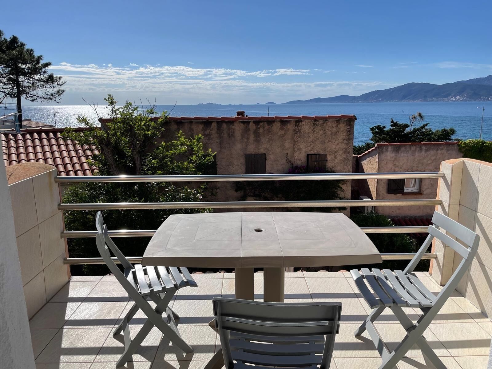 A balcony with a table and chairs, overlooking a coastal view with buildings and mountains in the distance.