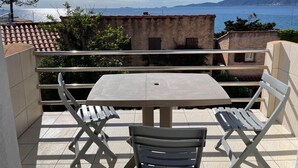 En balkon med et bord og stole, der giver udsigt over kysten med bygninger og bjerge i det fjerne.