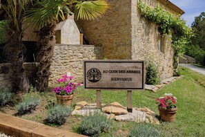 A stone building with a sign reading 'Au Coin des Arbres' and a welcome message. - Au Coin des Arbres - Genevrier (Biron)