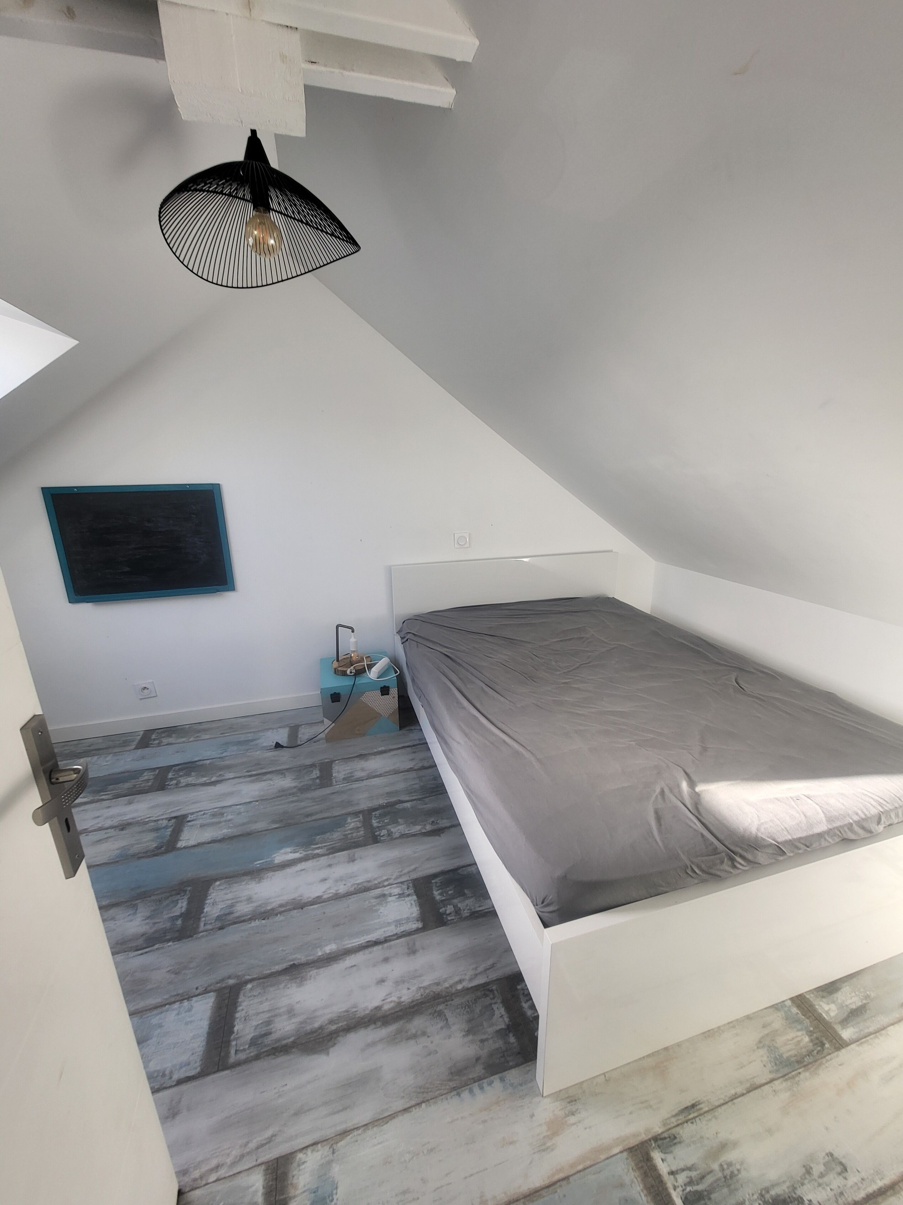 Um quarto pequeno com telhado inclinado, paredes brancas, uma cama de solteiro, luminária pendente em estrutura preta e uma pequena caixa azul com um objeto branco dentro.