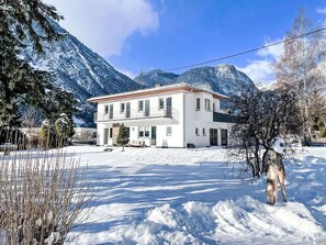Un edificio blanco de dos pisos con un balcón, rodeado de montañas y árboles cubiertos de nieve.