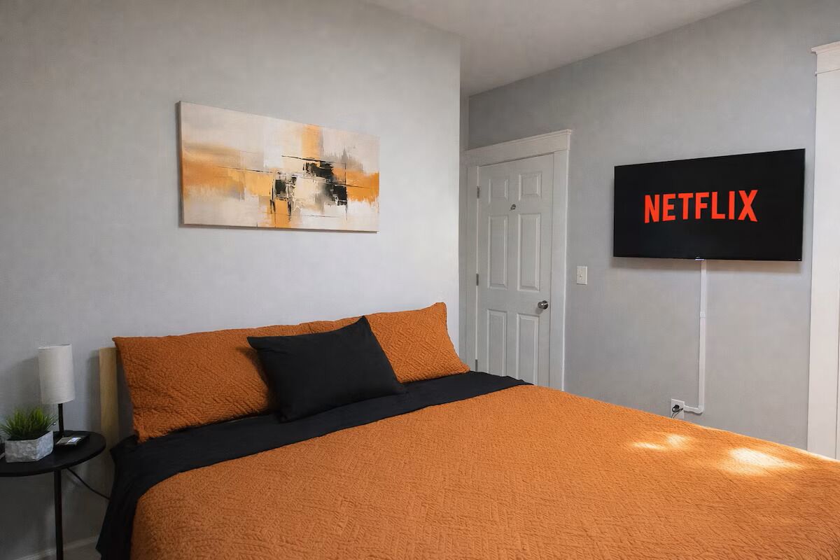 Ein Hotelzimmer mit einem Bett, einem an der Wand befestigten Fernseher, der "NETFLIX" zeigt, und einem Gemälde über dem Bett.