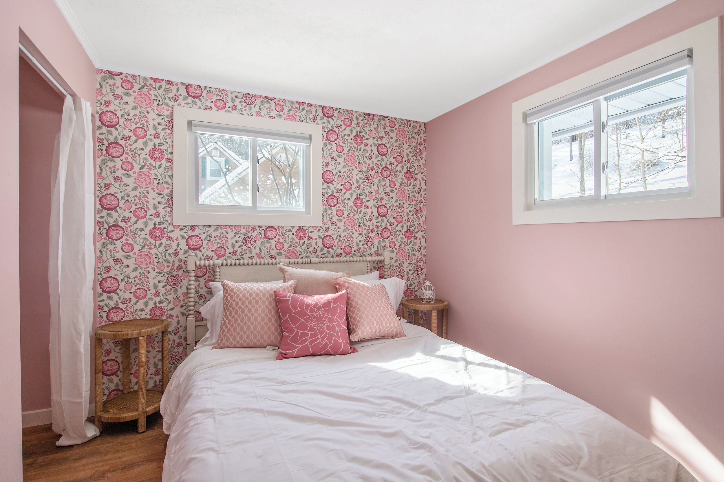 A bedroom with a floral wallpaper, a bed with white bedding and pink pillows, two bedside tables, and a window with a view of trees.