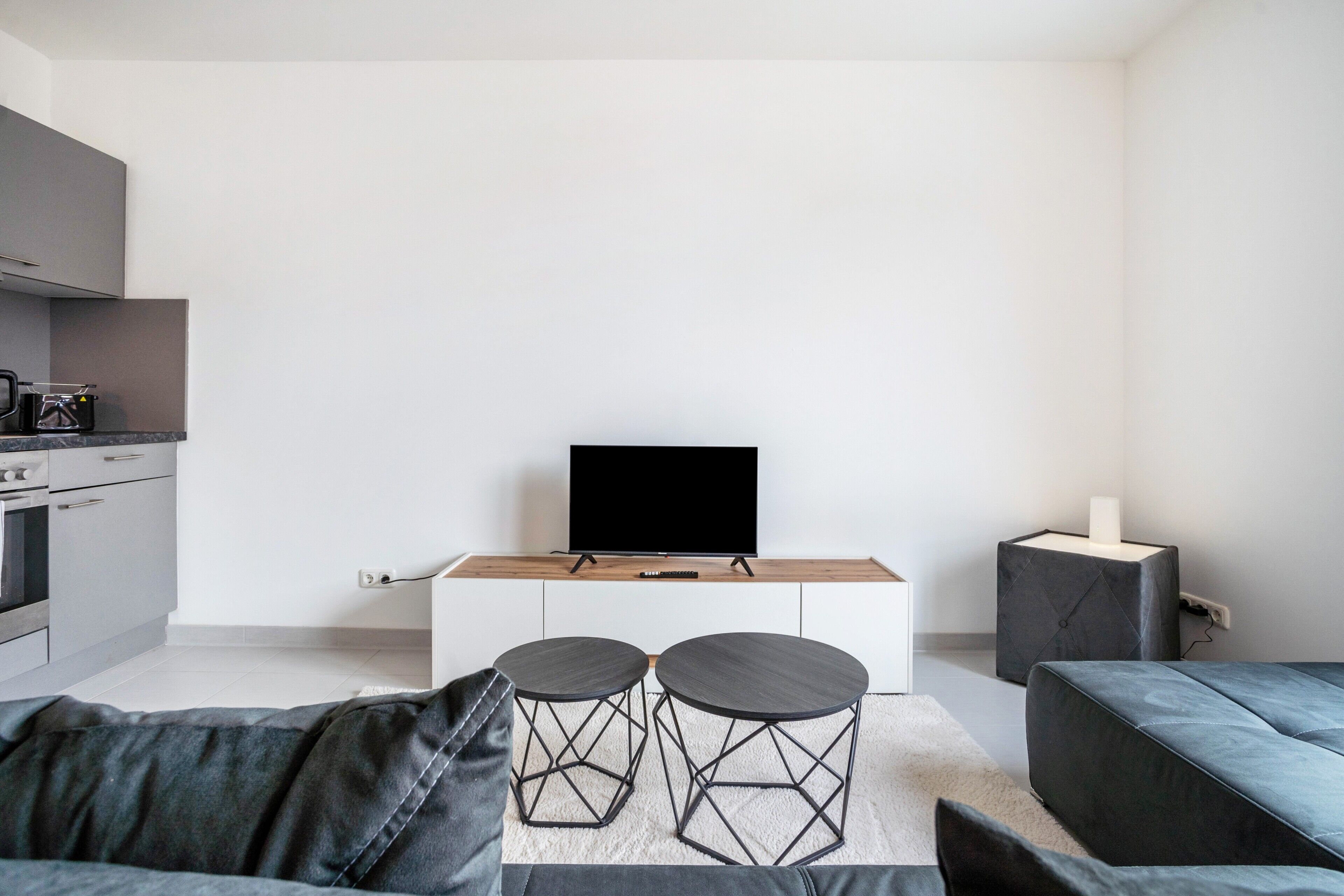 A modern living room with a grey sofa, two black stools, a flat-screen TV on a wooden stand, a grey cabinet, and a small side table with a candle.