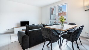 A modern living room with a sofa, dining table, chairs, a flat-screen TV, and a window with a view of buildings.