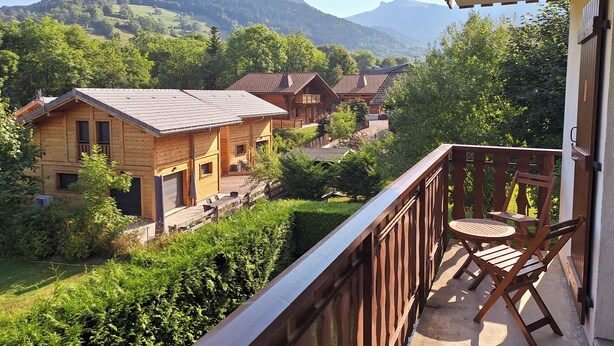 Een houten chalet met balkon, omgeven door weelderige begroeiing en bergen op de achtergrond.
