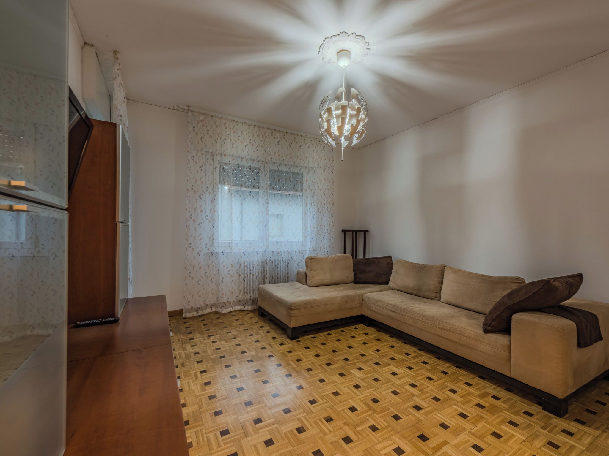 Un salon doté d’un parquet en bois, d’un canapé d’angle beige, d’une petite table et d’un luminaire décoratif.