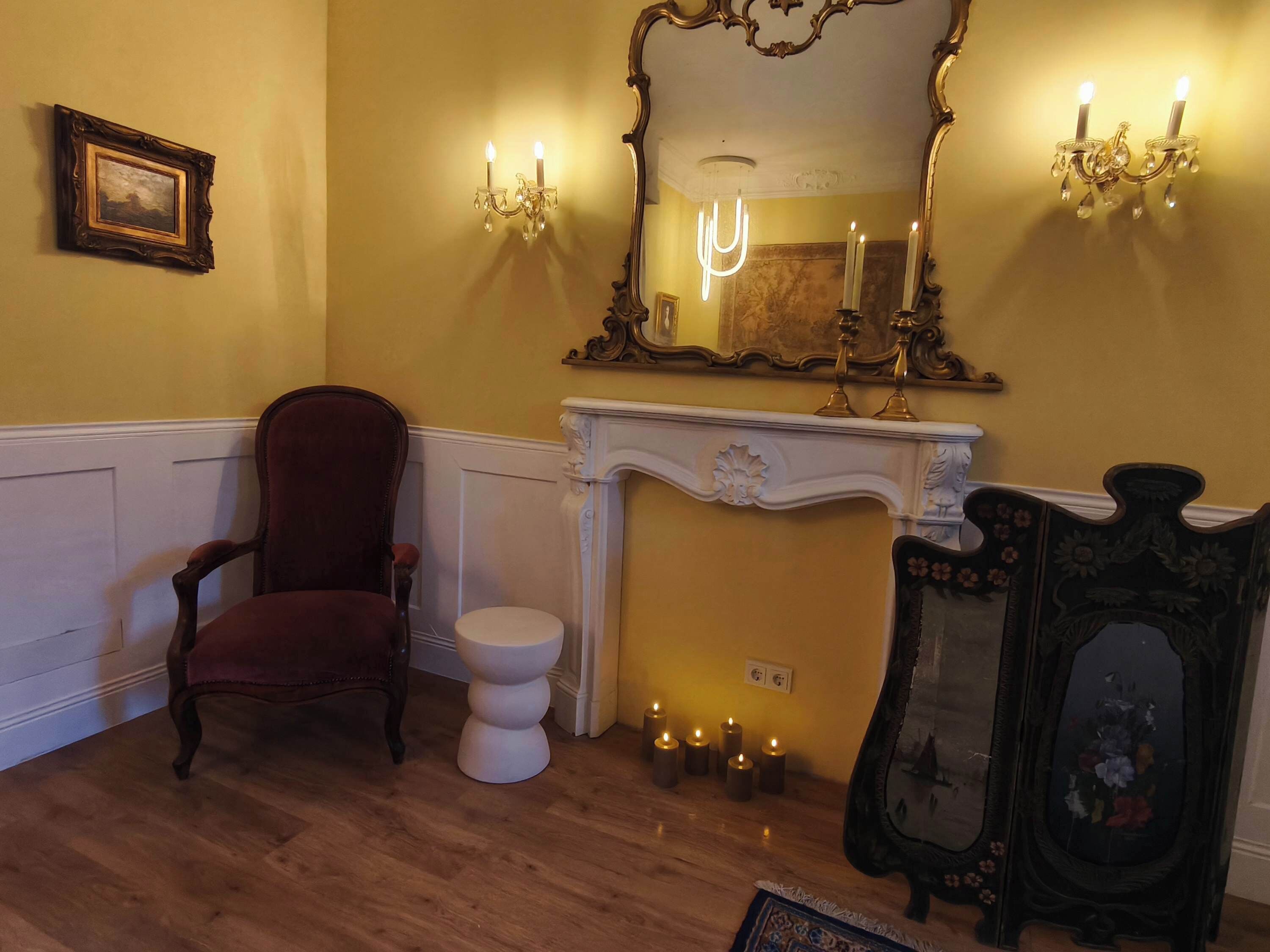 Una habitación con piso de madera, una silla roja, un inodoro blanco pedestal, una chimenea blanca con velas, un espejo grande con marco ornamentado y apliques de pared.