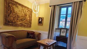 A living room with a wooden sofa, a small table, a large tapestry, and a window with curtains. - Elegance & Charm in Château – Suite Marie Thérèse (Malberg)