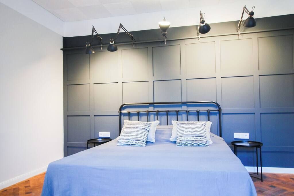 Un letto rifatto con cura, con copriletto blu e cuscini decorati, una testiera in metallo e tre lampade da lettura fissate alla parete.