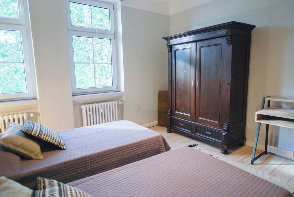 A bedroom with two beds, a large dark wooden wardrobe, a radiator, and a window with a view of greenery.