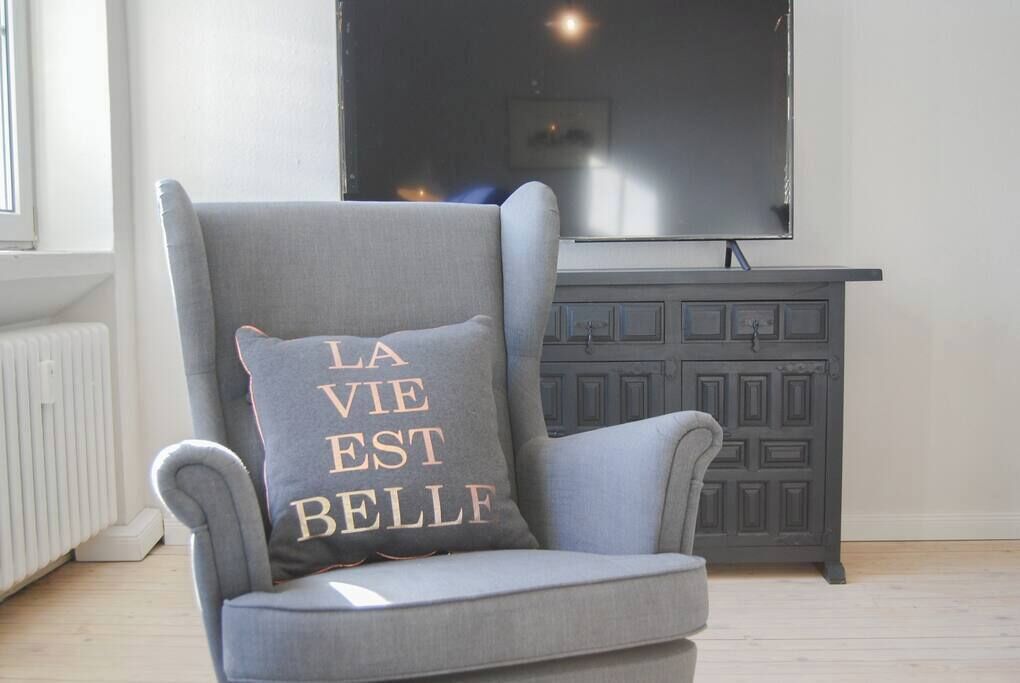 Een grijze fauteuil met een kussen waarop in gouden letters 'LA VIE EST BELLE' staat, geplaatst voor een donkergrijze kast en een grote flatscreen-televisie die aan de muur is bevestigd.