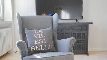 Een grijze fauteuil met een kussen waarop in gouden letters 'LA VIE EST BELLE' staat, geplaatst voor een donkergrijze kast en een grote flatscreen-televisie die aan de muur is bevestigd.