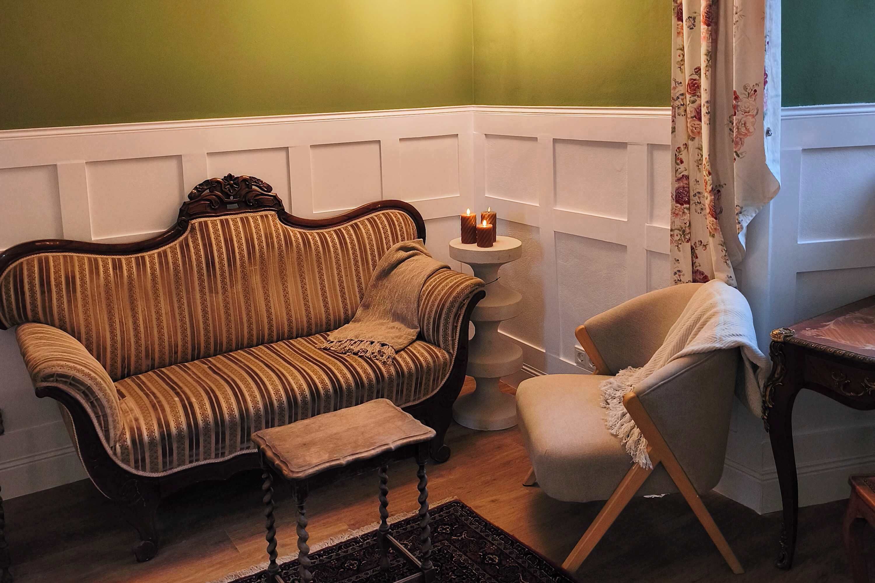 Ein Eckzimmer mit einem Vintage-Sofa mit Streifen, einem kleinen runden Tisch mit Kerzen und einem schlichten Sessel.