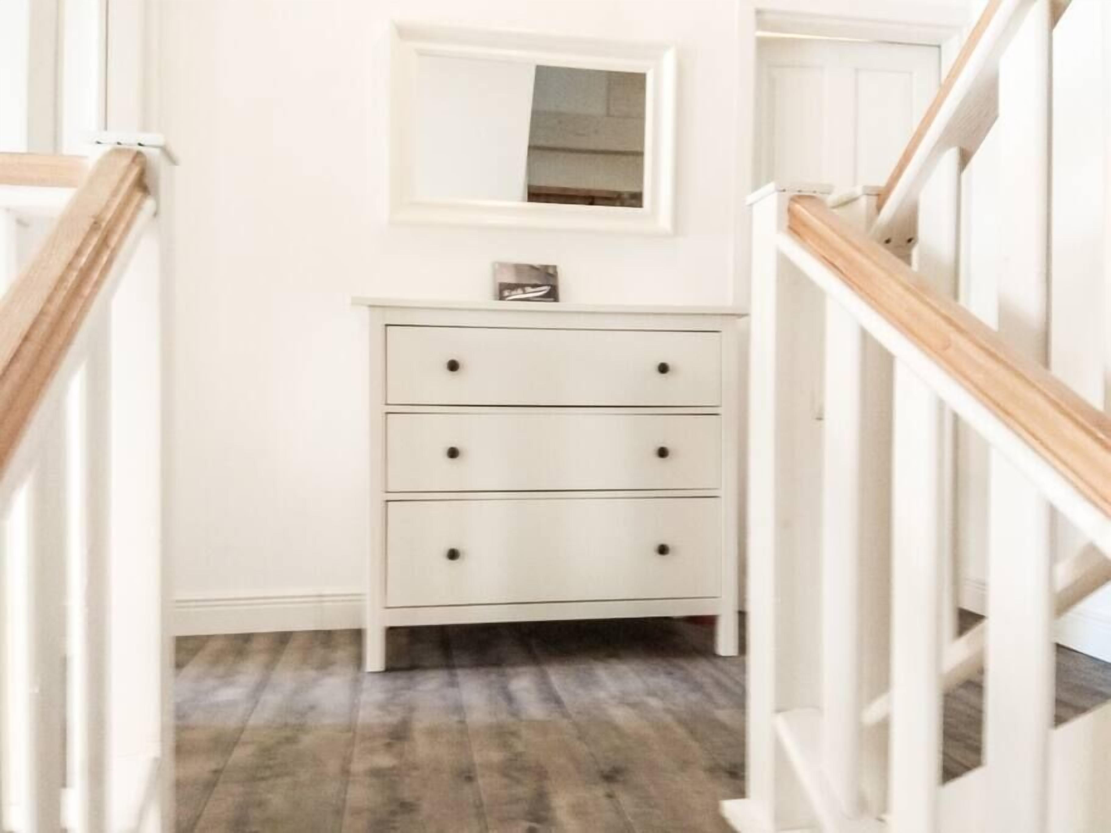 Un pasillo con un aparador blanco, un espejo pequeño encima y una escalera de madera a la derecha.