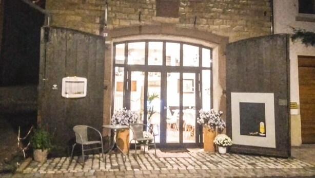 A stone building with a large arched entrance, outdoor seating area with metal chairs and tables, and potted plants. - Living in history: Heidi Braun Cottage (Dudeldorf)