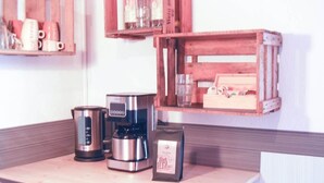 A kitchen counter with a coffee maker, a kettle, and a coffee bag. There are wooden shelves above the counter holding mugs and glasses.