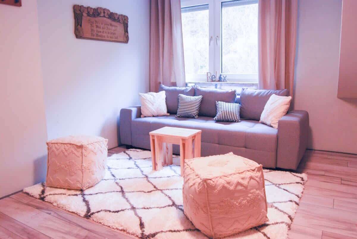 Een woonkamer met een grijze bank, twee voetenbankjes, een klein houten tafeltje en een kamerkleed met een patroon.
