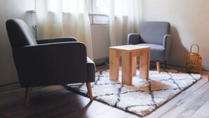 A room with a dark armchair, a grey armchair, a small wooden table, and a patterned rug on a tiled floor.