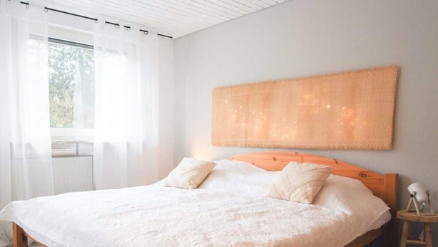 A bedroom with a large bed, white bedding, a wooden headboard, a bedside table with a lamp, and a window with sheer curtains.