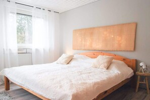 A bedroom with a large bed, white bedding, a wooden headboard, a bedside table with a lamp, and a window with sheer curtains.