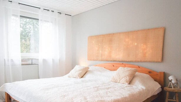 Un dormitorio con una cama grande, ropa de cama blanca, cabecera de madera, mesita de noche con lámpara y vistas a la vegetación a través de cortinas traslúcidas.