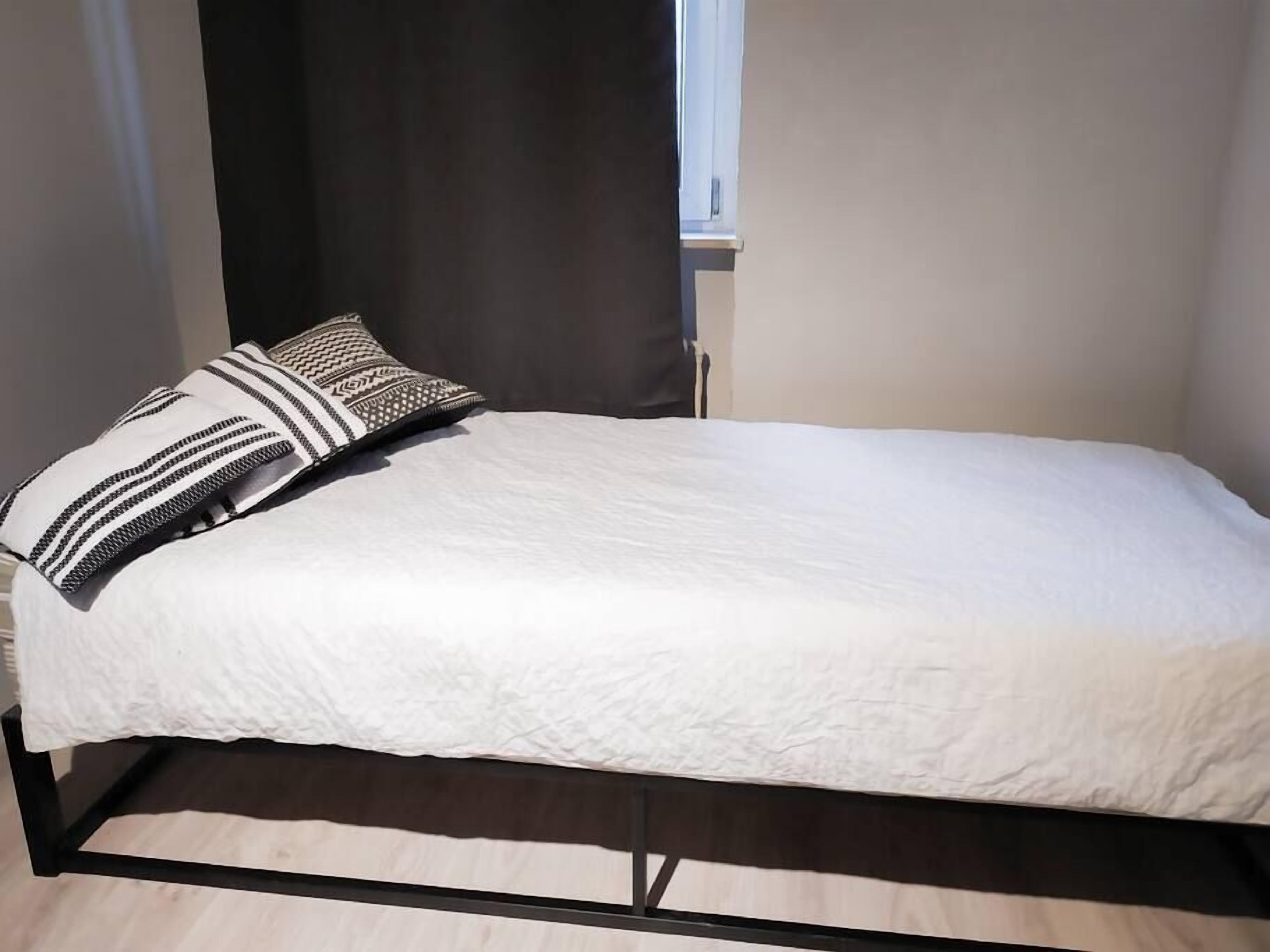Een enkel bed met een witte deken en twee gestreepte kussens, een donker metalen frame en een raam met gordijnen.