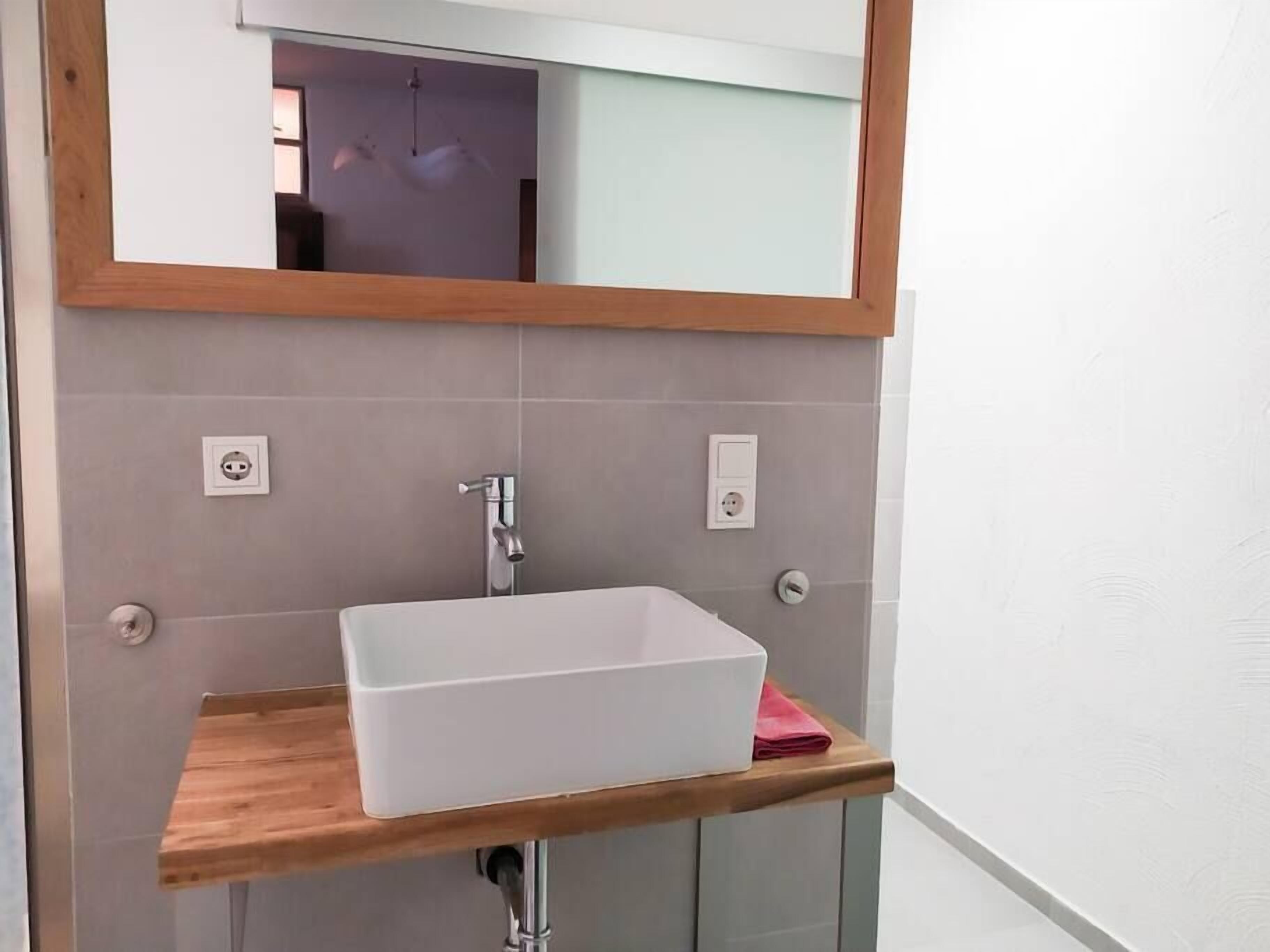 Un baño con un lavabo rectangular blanco, una encimera de madera, un espejo sobre el lavabo y una cabina de ducha con mampara de vidrio.