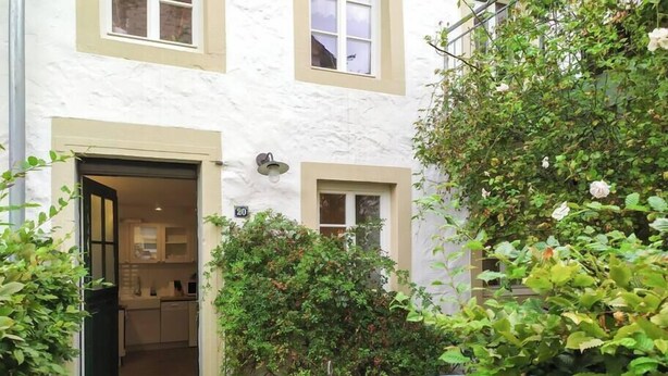 A white building with a door leading to a kitchen area, a small table, and a garden with various plants.