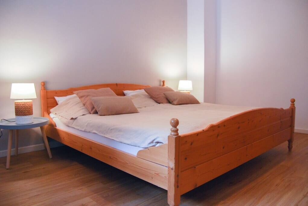 A wooden bed with a white mattress and beige pillows in a room with wooden flooring and white walls.