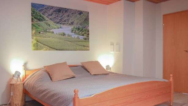 A bedroom with a wooden bed, two wall-mounted lamps, a painting of a landscape, and a wooden door. - Spacious room, private retro bathroom spring water (Traben-Trarbach)