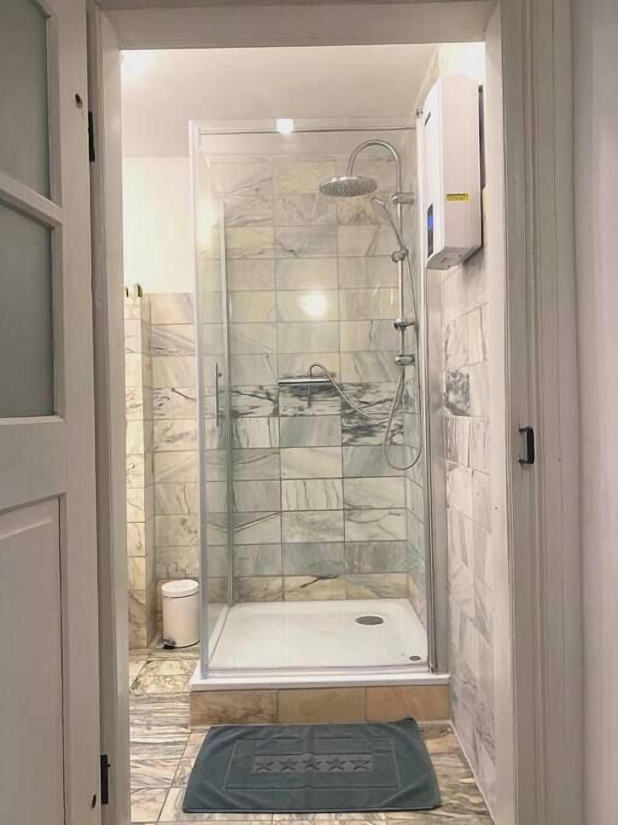 A glass-enclosed shower stall with a white floor, tiled walls, and a small white trash bin to the left.