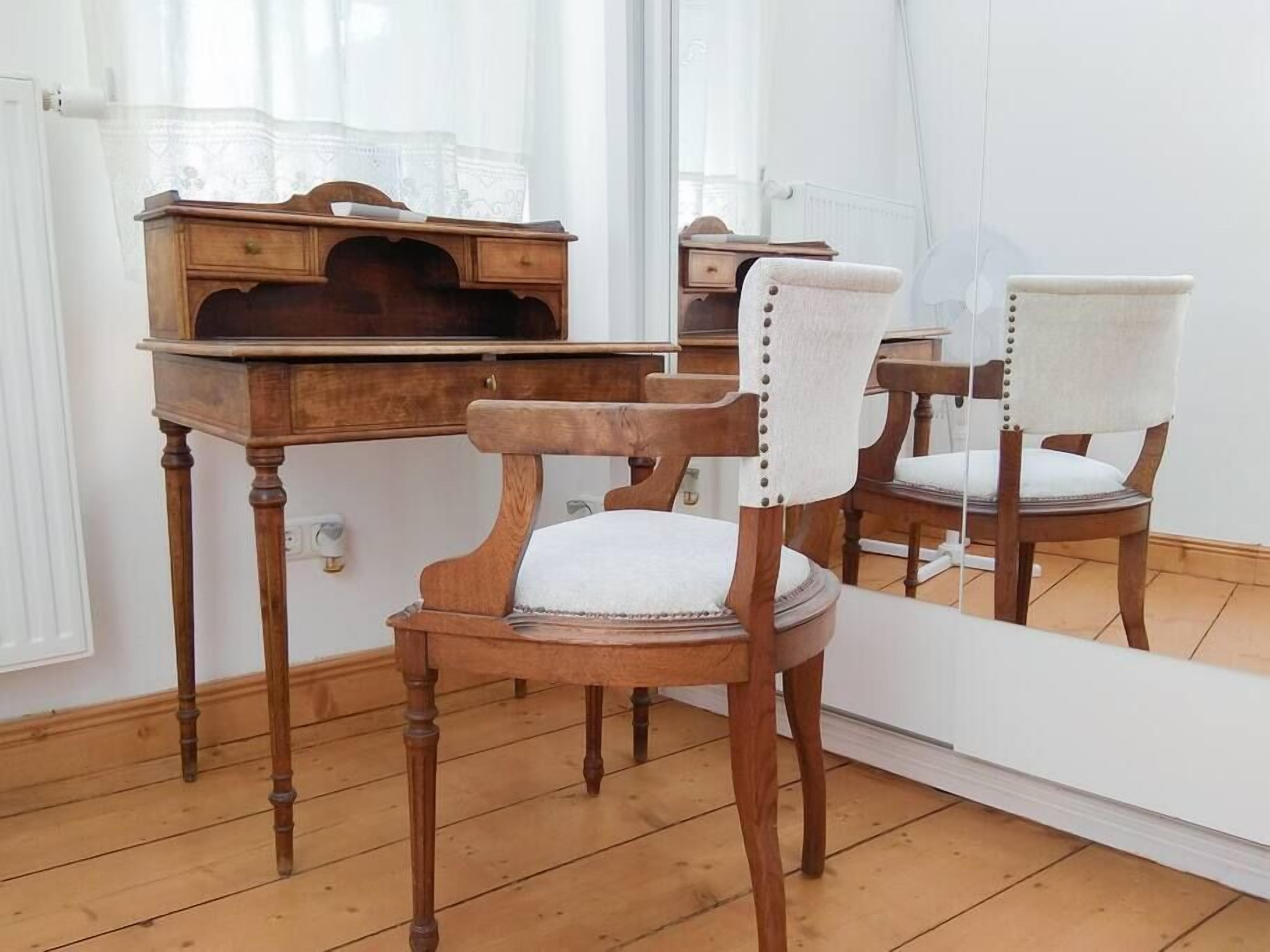 Een kamer met een houten bureau, een stoel met een witte kussen en een grote spiegel die het meubilair weerkaatst.