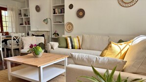Una sala de estar con sofá, mesa de centro y plantas en macetas. Se ve una estantería y un comedor al fondo.