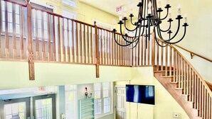 Un pasillo amplio con suelo de cuadros, una escalera de madera, un candelabro, un televisor montado en la pared y un sofá en la esquina.