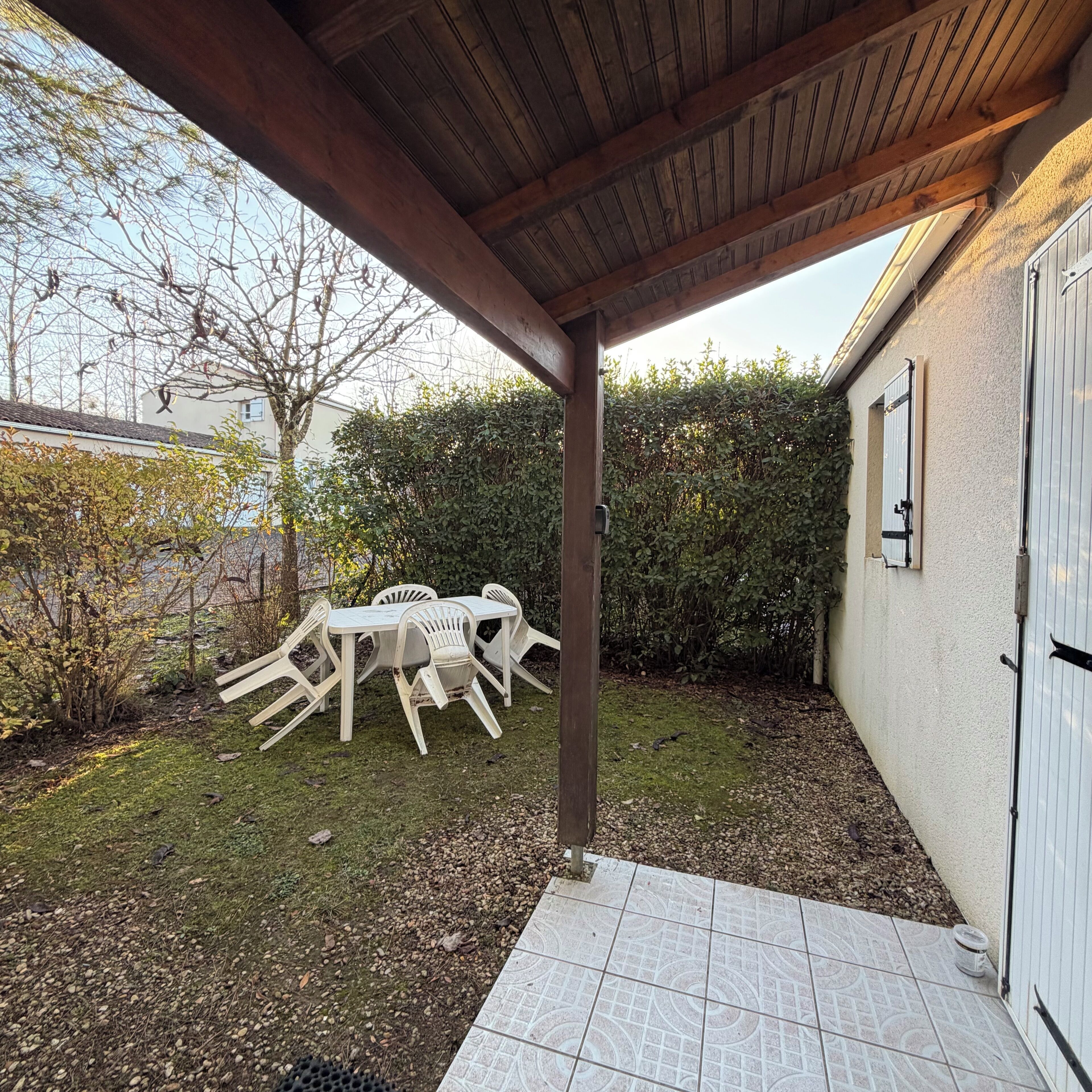 En overdækket terrasse med et bord og stole, flisegulv og en hæk i baggrunden.