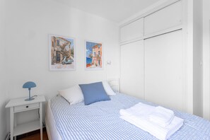 Quarto com uma cama, duas imagens penduradas na parede, uma mesa de cabeceira com abajur e um guarda-roupa.
