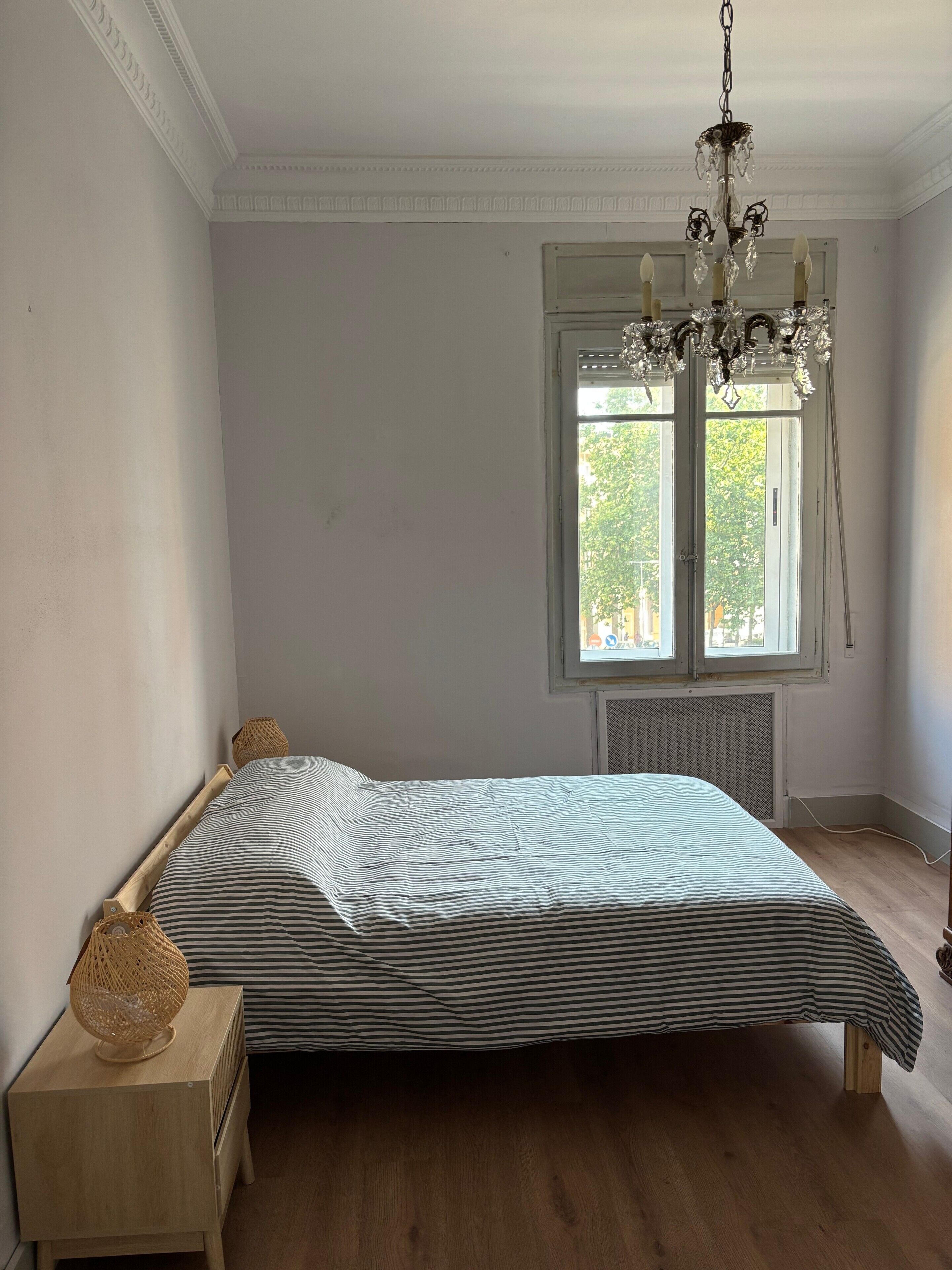 A bedroom with a bed, wooden nightstand, and a window with a view of trees.
