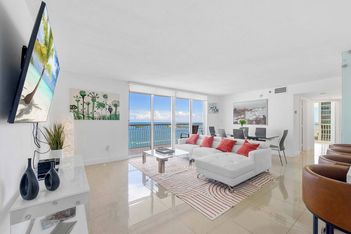 A modern living room with a white sofa, a coffee table, and a TV mounted on the wall. There is a large window with a view of the ocean.