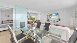 A modern living room with a glass dining table, white sofas, and a kitchen in the background. - The Club Luxury Condos by Ilios Rentals (Miami)
