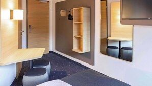 A modern hotel room with a wooden desk, a small seating area, a built-in shelf, and a flat-screen TV mounted on the wall. - B&B Hotel Rostock-Hbf (Rostock)