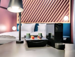 A neatly arranged hotel room with a modern bed, a bedside table with a lamp, a speaker, and a tray with cups and a bottle.