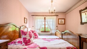 Un dormitori amb llit de fusta, una tauleta de nit, una commode, un penjallum i una finestra amb cortines.