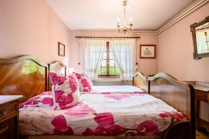 Ein Schlafzimmer mit einem hölzernen Bett, einem Nachttisch, einer Kommode, einer Chandelüster und einem Fenster mit Vorhängen.