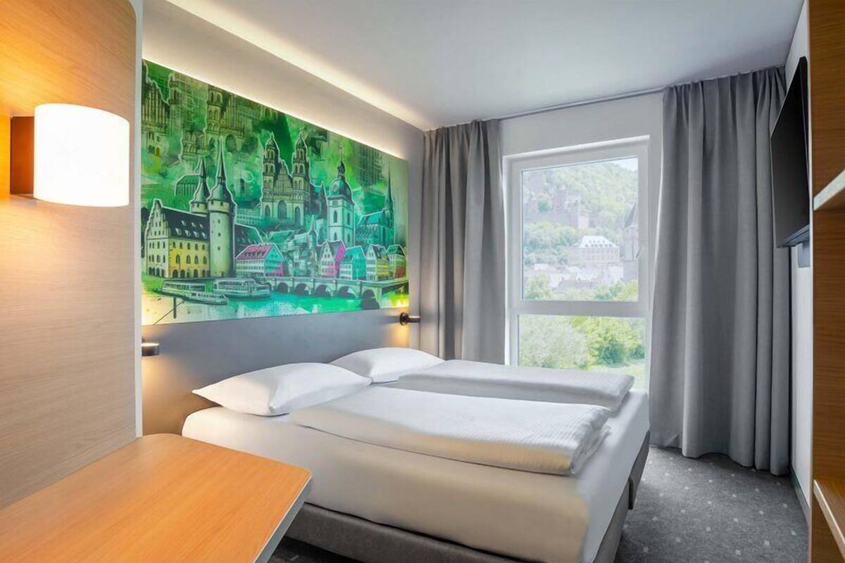 A hotel room with a bed, a desk, a painting of a cityscape, and a window with curtains.