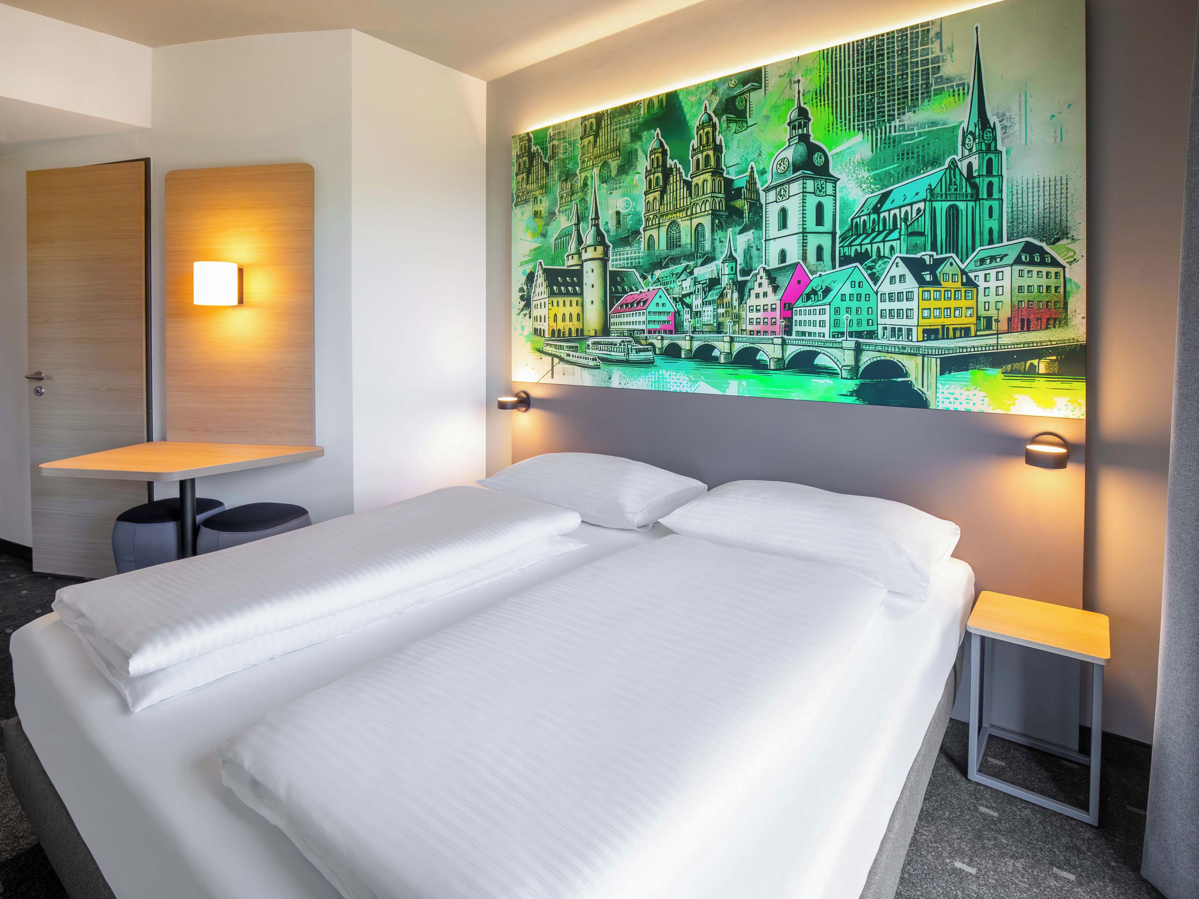 A hotel room with a bed, two bedside tables, a wall-mounted lamp, and a colorful cityscape mural.
