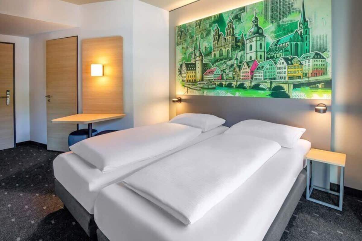 A hotel room with a bed, two bedside tables, a wall-mounted light, and a large cityscape painting.