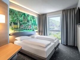 Habitación de hotel con dos camas, un cuadro de un paisaje urbano, un escritorio y una ventana con cortinas.