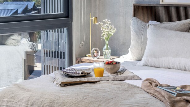 A modern bedroom with a large window, a bedside table with a vase of flowers, a bowl of fruit, and a glass of orange juice. - Pueblo La Dehesa (Lo Barnechea)