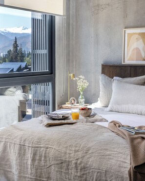 Ein modernes Schlafzimmer mit großem Fenster, einem Nachttisch mit Blumenvase, Obstschale und Glas Orangensaft.